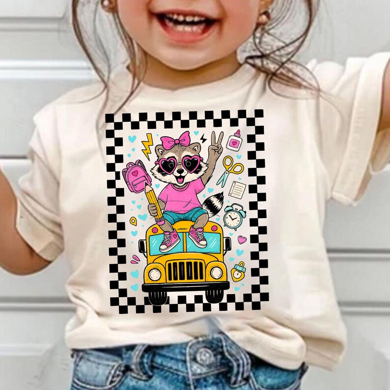 May include: A cream-colored t-shirt featuring a cartoon raccoon wearing sunglasses and a pink bow, holding a pencil and making a peace sign. The raccoon is riding a yellow school bus, surrounded by school-themed graphics.