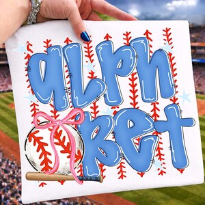 May include: White square with the word "ALPHABET" in large, light blue bubble letters. A baseball with a pink bow and a small wooden bat are featured below the text. Red stitching and blue stars decorate the background.