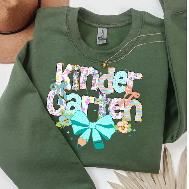 May include: An olive green sweatshirt with the words "Kindergarten" in a colorful, floral design. The design includes a turquoise bow and pencil, with additional floral accents. A gold necklace is visible.