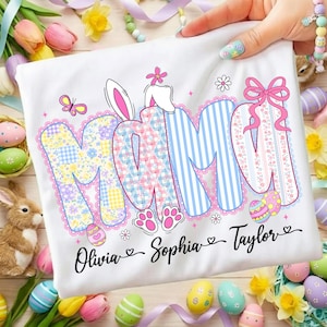 May include: White t-shirt with the word "MOM" in colorful, patterned letters, featuring a bunny ear design. The names "Olivia", "Sophia", and "Taylor" are written below. Easter-themed with eggs and flowers.