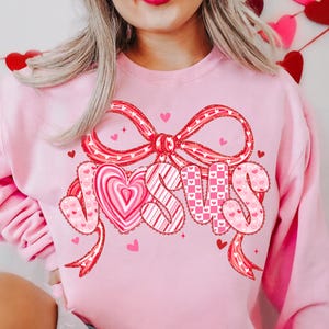May include: A light pink sweatshirt with a large bow and the word "JESUS" in a heart-themed design. The letters are decorated with hearts and patterns in red and pink, perfect for Valentine's Day or religious celebrations.