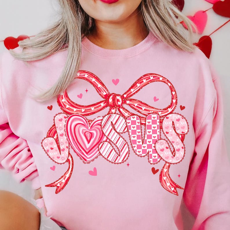 May include: A light pink sweatshirt with a large bow and the word "JESUS" in a heart-themed design. The letters are decorated with hearts and patterns in red and pink, perfect for Valentine's Day or religious celebrations.