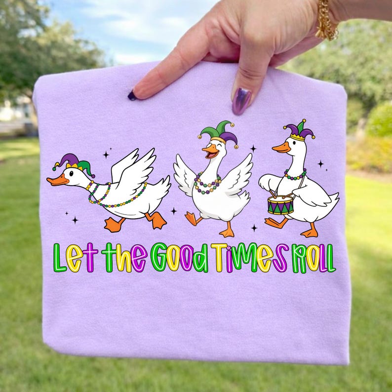 May include: A light purple t-shirt featuring three cartoon geese in Mardi Gras attire. The geese are wearing jester hats and beaded necklaces, with one playing a drum. The text "Let the Good Times Roll" is below the geese.