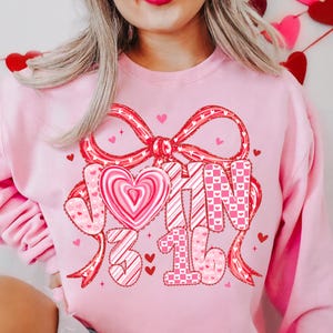 May include: A light pink sweatshirt with a large red bow and the words "JOHN 3:16" in a decorative font. The design includes a heart-shaped candy and small hearts scattered around the text, creating a Valentine's Day theme.
