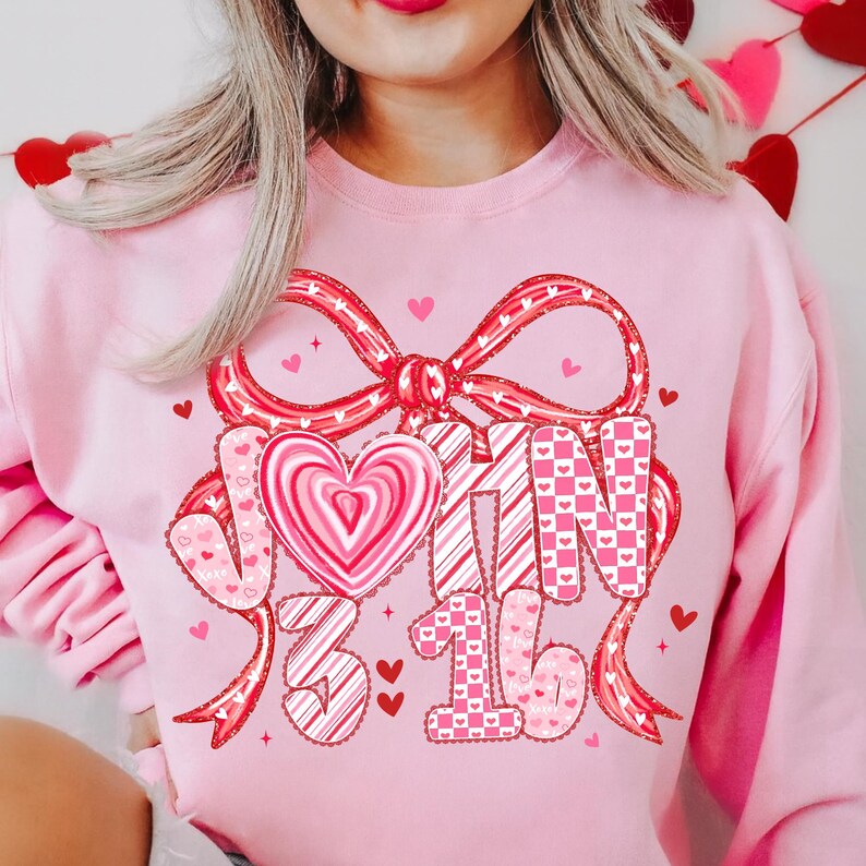 May include: A light pink sweatshirt with a large red bow and the words "JOHN 3:16" in a decorative font. The design includes a heart-shaped candy and small hearts scattered around the text, creating a Valentine's Day theme.