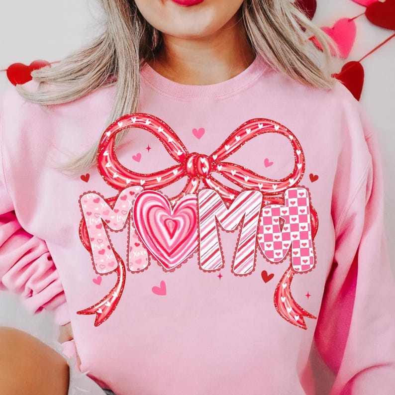 May include: Pink sweatshirt with a large red bow and the word "MOM" in a decorative font. The letters are adorned with hearts and patterns. Small red hearts are scattered around the design, creating a festive look.
