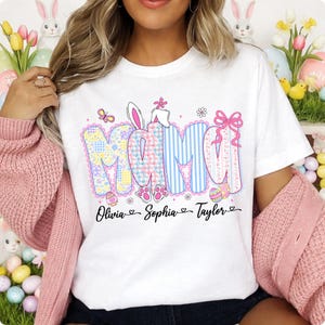 May include: White t-shirt with the word "MAMA" in pastel colors and Easter-themed designs. The shirt features the names "Olivia", "Sophia", and "Taylor" below the word "MAMA". The design includes bunny ears, eggs, and paw prints.