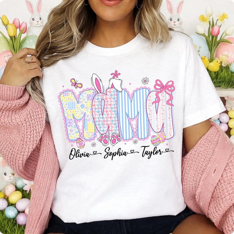May include: White t-shirt with the word "MAMA" in pastel colors and Easter-themed designs. The shirt features the names "Olivia", "Sophia", and "Taylor" below the word "MAMA". The design includes bunny ears, eggs, and paw prints.