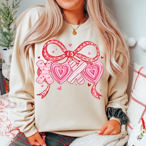 May include: A cream-coloured sweatshirt with a Valentine's Day design. The graphic features a large red bow, pink hearts, and the letters "XOX" in a heart shape. Small pink hearts are scattered around the design.
