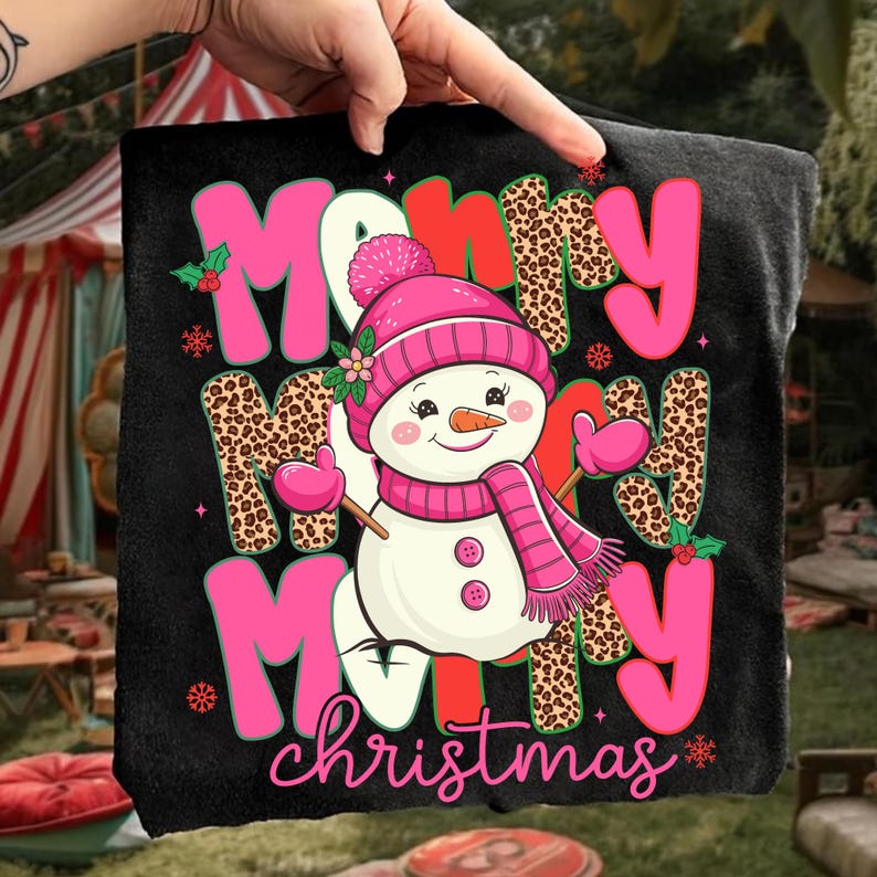 May include: Black t-shirt with a festive design featuring a snowman wearing a pink hat, scarf, and mittens. The words "Merry Christmas" are displayed in pink and leopard print lettering, with holly and snowflake accents.