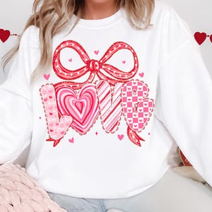 May include: White sweatshirt with a large red bow and the word "LOVE" in pink and red lettering. The "O" is a heart shape, and the design includes small hearts and decorative accents, perfect for Valentine's Day.