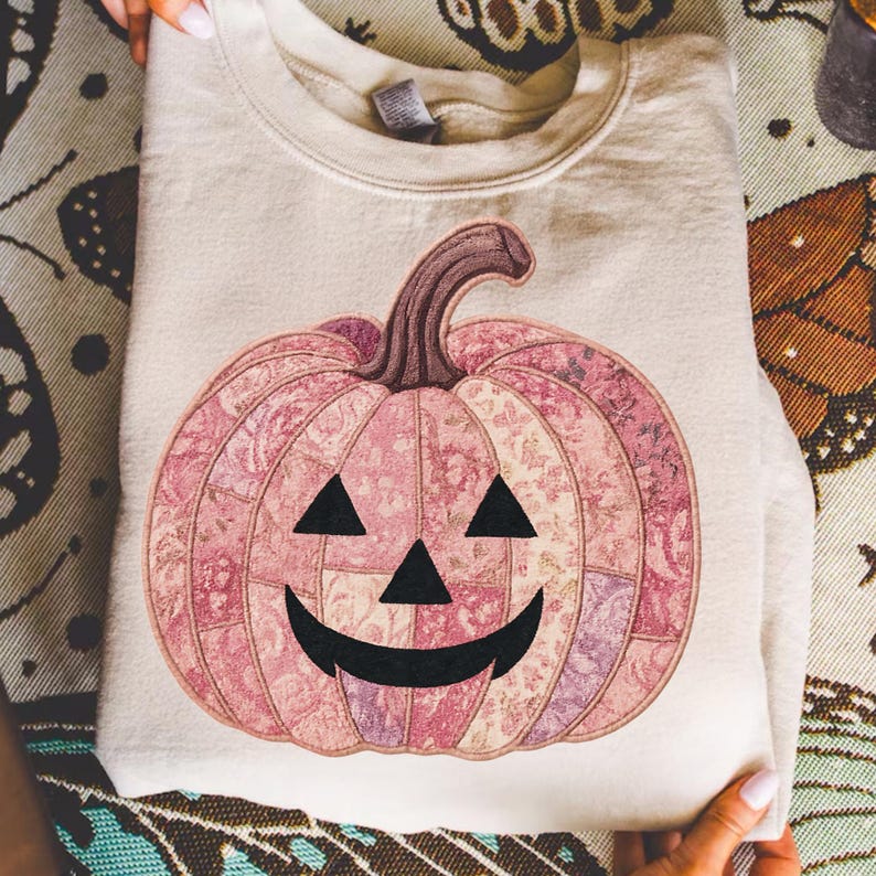 K&ouml;nnte beinhalten: Ein cremefarbenes Sweatshirt mit einem rosa K&uuml;rbisdesign. Der K&uuml;rbis hat ein Patchworkmuster mit floralen Details, einen braunen Stiel und ein klassisches Jack-o'-Lantern-Gesicht mit schwarzen Dreiecksaugen und einem gebogenen Mund. Ein festliches Kleidungsst&uuml;ck f&uuml;r Halloween.