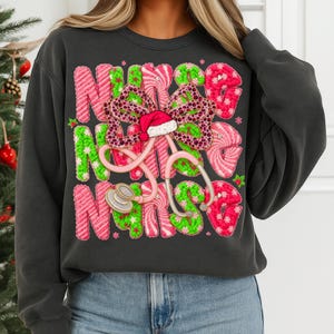 May include: Dark gray sweatshirt with the word "NURSE" in pink and green candy cane-style letters. A stethoscope and a bow with a Santa hat are in the center. The sweatshirt is a festive holiday design.