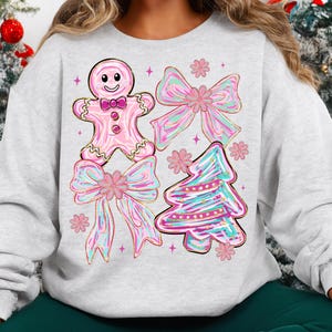 May include: A light grey sweatshirt with a festive design. The design features a pink gingerbread man, two bows, and a Christmas tree, all with pink, blue, and gold accents. The sweatshirt is a comfortable, casual style.