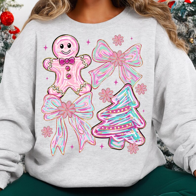 May include: A light grey sweatshirt with a festive design. The design features a pink gingerbread man, two bows, and a Christmas tree, all with pink, blue, and gold accents. The sweatshirt is a comfortable, casual style.
