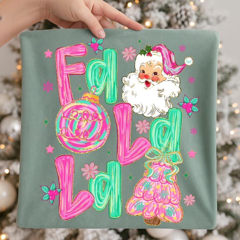 May include: A decorative pillow with a festive design. The pillow features the words "Fa La La" in pink and green, along with a Santa Claus illustration and Christmas tree. The design includes a Christmas ornament and floral accents.