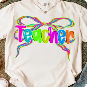 Op de afbeelding: Cr&egrave;mekleurig T-shirt met het woord "Teacher" in levendige, veelkleurige bubbelletters. Een kleurrijk strikdesign, met neon tinten roze, groen, blauw en oranje, omlijst het woord. Het shirt is gemaakt van een zacht materiaal.