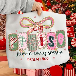 May include: White sweatshirt with the text "I will praise Him in every season" and Psalm 146:2. The word "praise" is in colorful, patterned letters. A pink and gold bow is at the top.