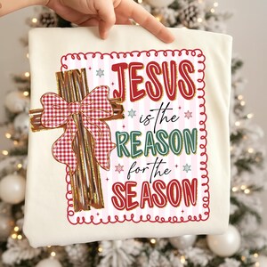 May include: Cream-colored t-shirt featuring a decorative cross with a red and white plaid bow. The shirt has the text "JESUS is the REASON for the SEASON" in red, green, and blue, with snowflake accents, perfect for Christmas.