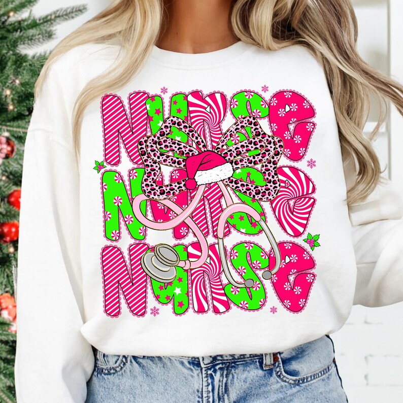 May include: White sweatshirt with a festive design. The design features the words "NURSE" in pink and green, a leopard print bow, a Santa hat, and a stethoscope. The design is decorated with snowflakes and stars.