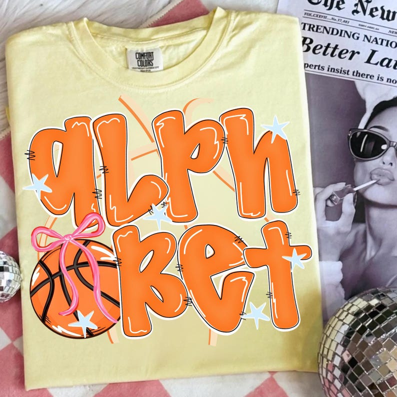 May include: A light yellow t-shirt with the word "ALPHABET" in orange bubble letters. A basketball with a pink bow is on the left side. The shirt has a crew neck and short sleeves. The text is outlined in white.