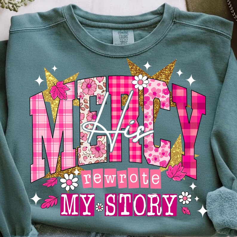 Op de afbeelding: Teal sweatshirt met de tekst "MERCY His rewrote MY STORY" in roze, bloemen- en ruitpatronen, geaccentueerd met gouden glittersterren en witte madeliefjes. Een casual top.