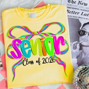 May include: A yellow t-shirt featuring the word "Senior" in large, colourful letters, with "Class of 2026" underneath. The design includes a rainbow-coloured bow, creating a celebratory and eye-catching look.