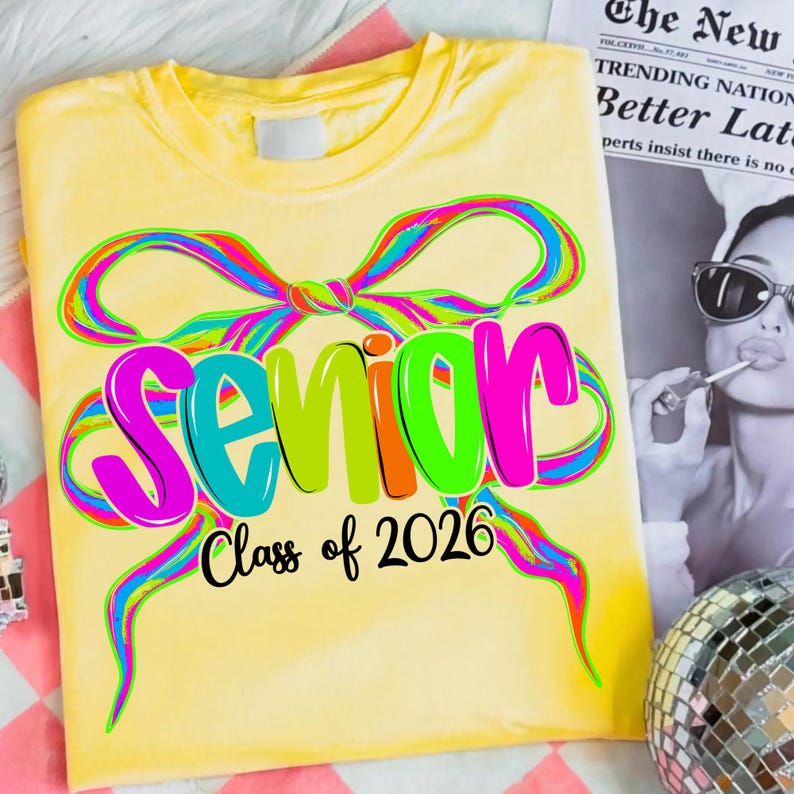 May include: A yellow t-shirt featuring the word "Senior" in large, colourful letters, with "Class of 2026" underneath. The design includes a rainbow-coloured bow, creating a celebratory and eye-catching look.