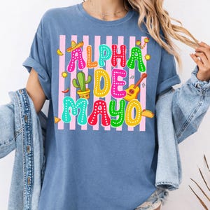 May include: A blue t-shirt with the words "ALPHA DE MAYO" in colorful letters. The design includes a sombrero, cactus, guitar, and other festive elements, set against a pink and white striped background.