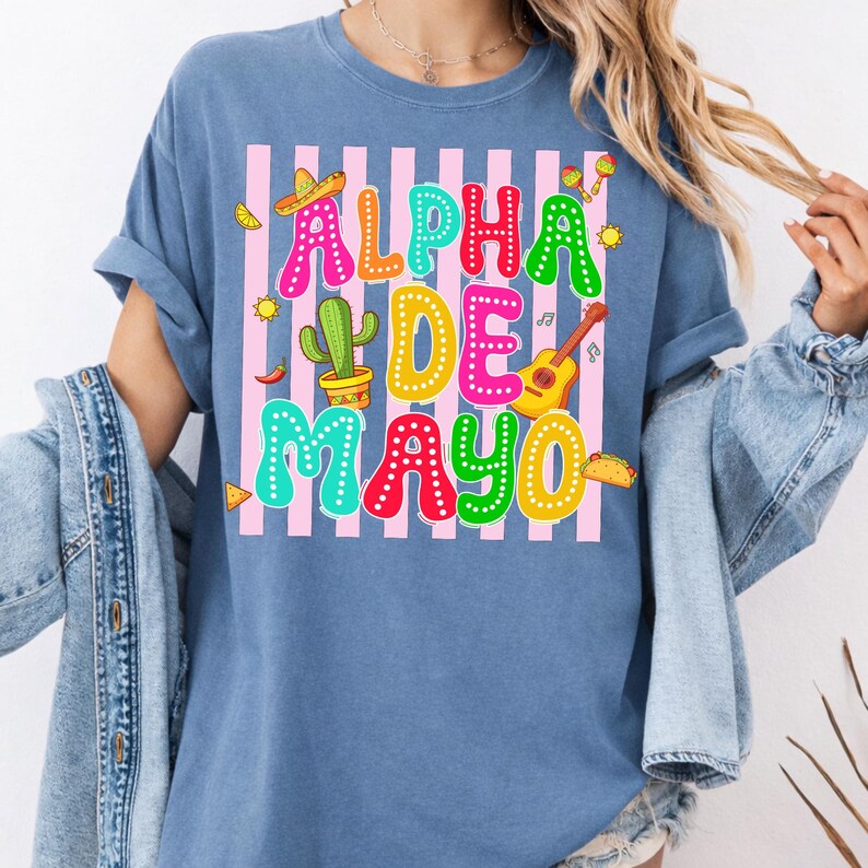 May include: A blue t-shirt with the words "ALPHA DE MAYO" in colorful letters. The design includes a sombrero, cactus, guitar, and other festive elements, set against a pink and white striped background.