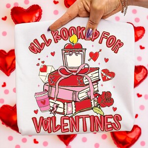 May include: White sweatshirt with a Valentine's Day graphic. The design features a stack of books, a candle, a coffee cup, and a rose. The text reads "ALL BOOKED UP FOR VALENTINES" in red.