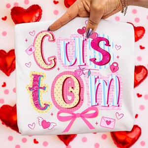 May include: White t-shirt with the word "CUSTOM" in a colorful, decorative font. The letters are pink, blue, and yellow, with heart and rose accents. A pink bow and heart-shaped balloons are also visible.