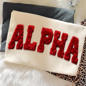May include: Cream-colored pillow with the word "ALPHA" in bold, red, fuzzy letters. The pillow is rectangular and rests on a dark gray pillow and a white, fluffy surface. A leopard print fabric is visible on the right.