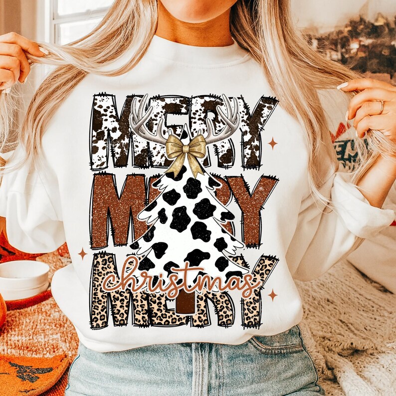 May include: White sweatshirt with a festive design. The design features the words "Merry Christmas" in a mix of fonts and patterns, including a cow print Christmas tree, antlers, and a gold bow. The overall theme is holiday apparel.