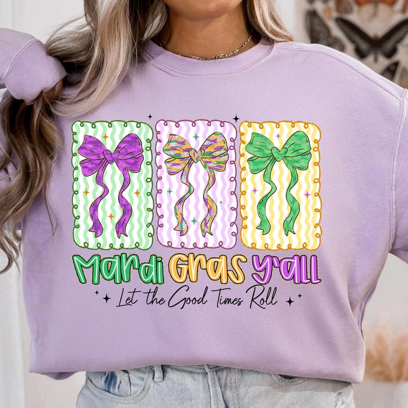 May include: A light purple sweatshirt with a graphic design featuring three colorful bows in purple, gold, and green. The text "Mardi Gras y'all" and "Let the Good Times Roll" are printed below the bows.