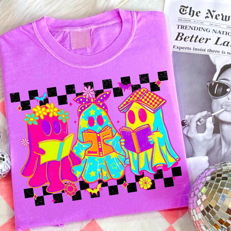 May include: A lilac-colored t-shirt featuring a vibrant graphic of three cartoon ghosts reading books. The ghosts are in bright colors with floral and star accents, set against a black and white checkered pattern. The text on the newspaper is blurry.