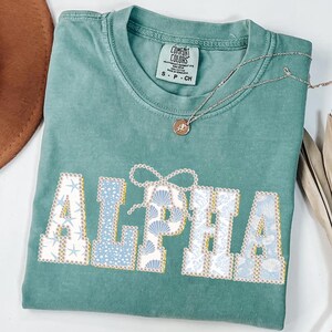 May include: A seafoam green Comfort Colors t-shirt with the word "ALPHA" in a decorative font. The letters are filled with seashell and starfish patterns in shades of blue and white. A silver necklace with a small pendant rests on the shirt.
