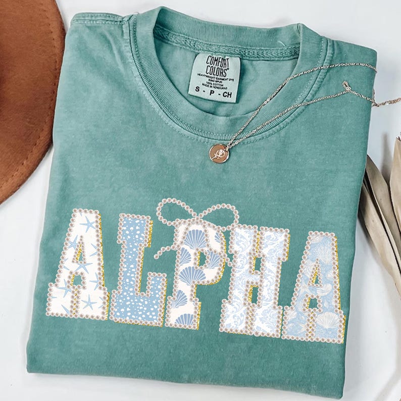 May include: A seafoam green Comfort Colors t-shirt with the word "ALPHA" in a decorative font. The letters are filled with seashell and starfish patterns in shades of blue and white. A silver necklace with a small pendant rests on the shirt.