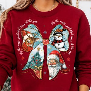 May include: A red sweatshirt featuring a festive bow design with a reindeer, snowman, Christmas tree, and Santa Claus. The words "It's the most wonderful time of the year" are written around the bow in a circular pattern.