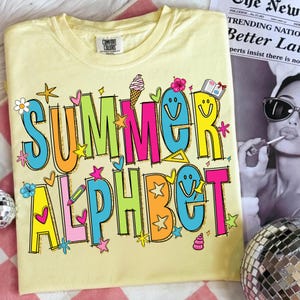 May include: A pale yellow t-shirt featuring the words "SUMMER ALPHABET" in a vibrant, cartoonish font. The design incorporates stars, flowers, and ice cream motifs. The shirt has a round neck and short sleeves.