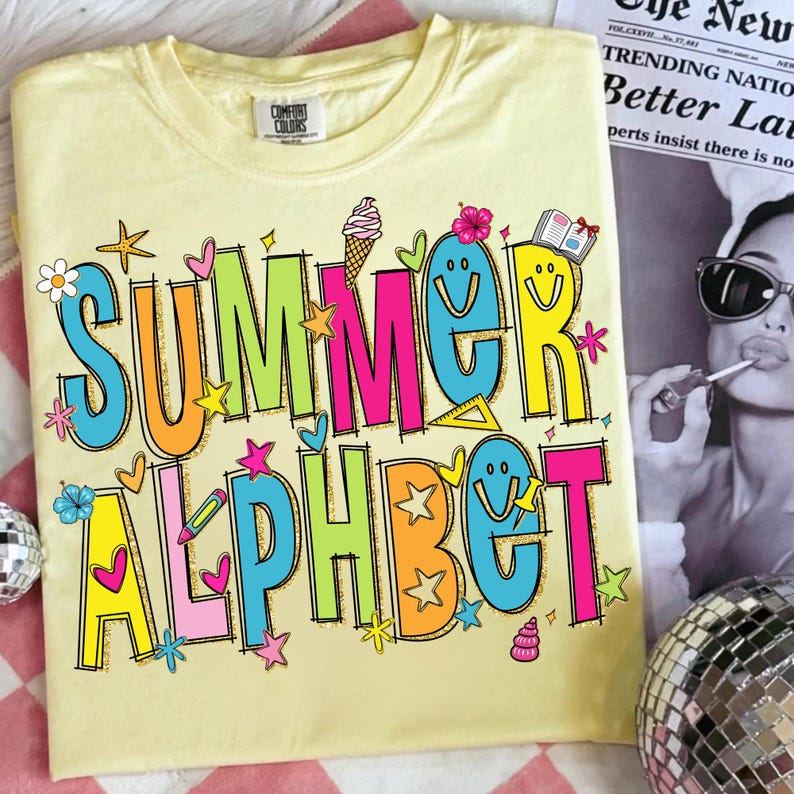 May include: A pale yellow t-shirt featuring the words "SUMMER ALPHABET" in a vibrant, cartoonish font. The design incorporates stars, flowers, and ice cream motifs. The shirt has a round neck and short sleeves.
