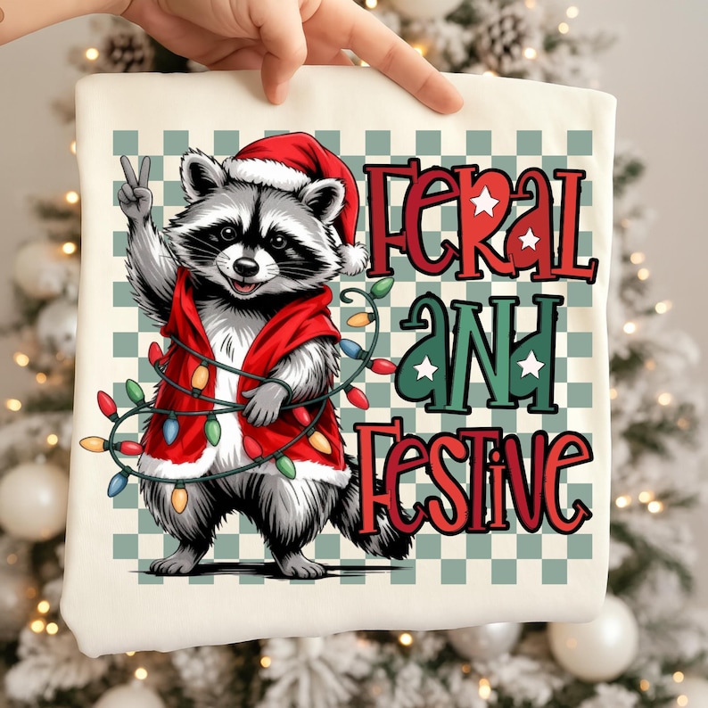 May include: A cream-colored tote bag featuring a raccoon wearing a Santa hat and red vest, holding a string of Christmas lights. The text "FERAL AND FESTIVE" is displayed in red and green. The background has a green and white checkered pattern.