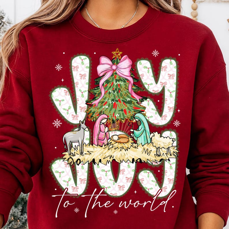 May include: A maroon sweatshirt with a Christmas-themed graphic. The design features the word "JOY" in large, decorative letters, a Christmas tree, and a nativity scene. The phrase "to the world" is written below the word "JOY".