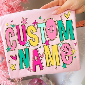 May include: A light pink t-shirt with the words "CUSTOM NAME" in a colorful, playful design. The text is outlined in black and features pink, yellow, teal, and green colors, with decorative stars, hearts, and smiley faces.
