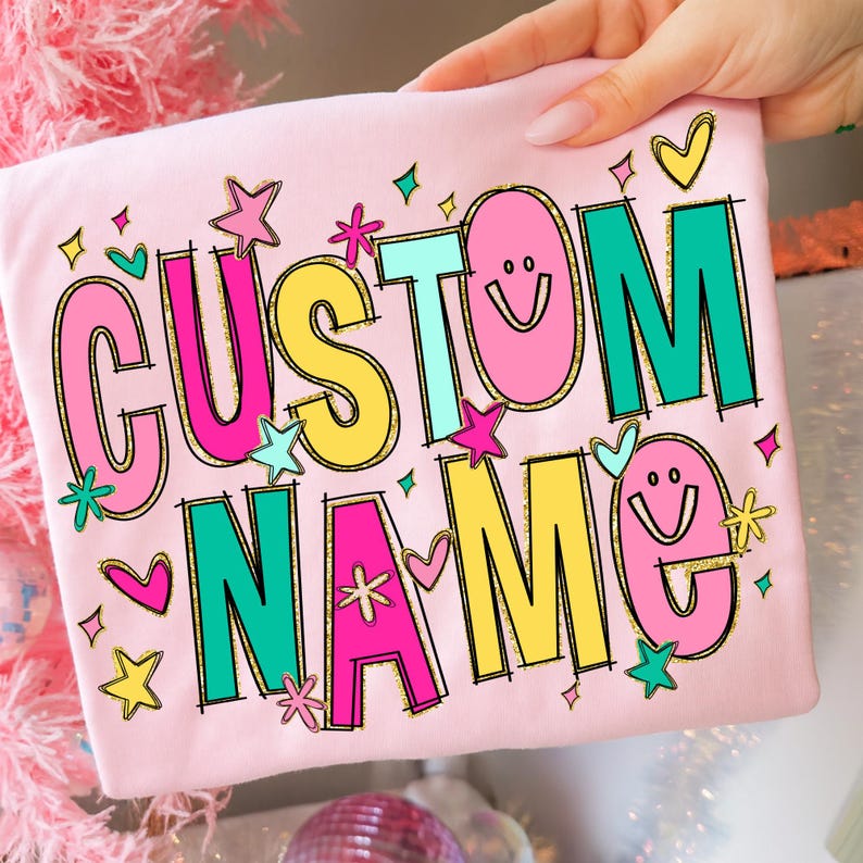 May include: A light pink t-shirt with the words "CUSTOM NAME" in a colorful, playful design. The text is outlined in black and features pink, yellow, teal, and green colors, with decorative stars, hearts, and smiley faces.