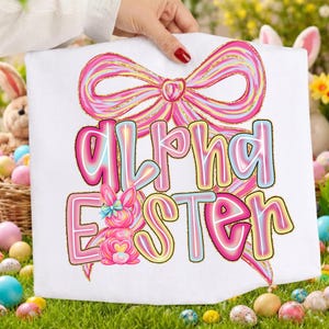 May include: White t-shirt featuring a large pink bow and the words "Alpha Easter" in a colorful, outlined font. A pink bunny with a blue bow is also featured. The design is set against a background of Easter eggs and grass.