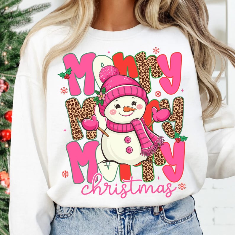 May include: White sweatshirt with a festive design. The design features the words "Merry Christmas" in pink and leopard print, along with a snowman wearing a pink hat, scarf, and mittens. Snowflakes and holly accents complete the holiday theme.
