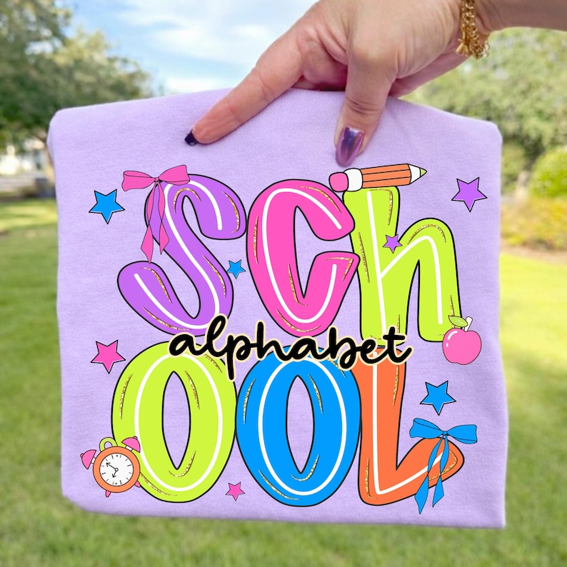 May include: A lilac t-shirt featuring the words "SCHOOL ALPHABET" in a vibrant, multi-coloured design. The graphic includes a pencil, apple, stars, and a clock. The letters are outlined in pink, green, blue, and orange.