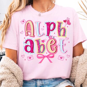 May include: A light pink t-shirt with the word "ALPHABET" in colorful, decorative letters. The letters are in various patterns and colors, with hearts and bows. The shirt is a Valentine's Day theme.
