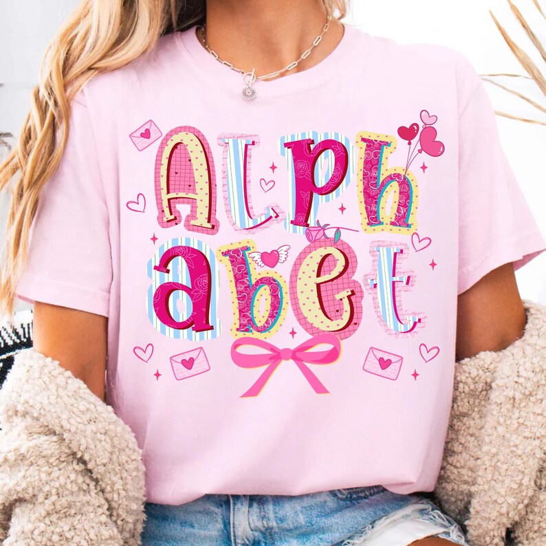 May include: A light pink t-shirt with the word "ALPHABET" in colorful, decorative letters. The letters are in various patterns and colors, with hearts and bows. The shirt is a Valentine's Day theme.